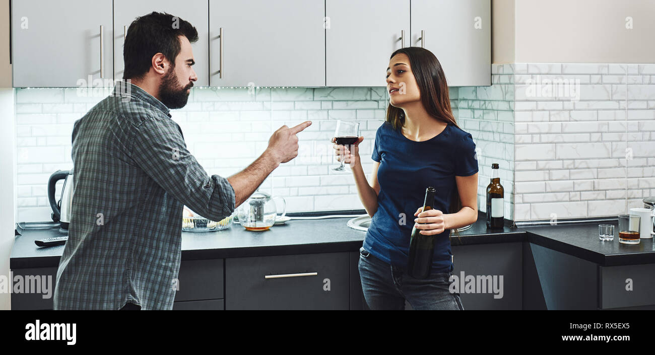 Drunkard. Dark-haired addicted woman arguing with her husband. She is standing in the kitchen with a glass and a bottle of red wine and listening to h Stock Photo