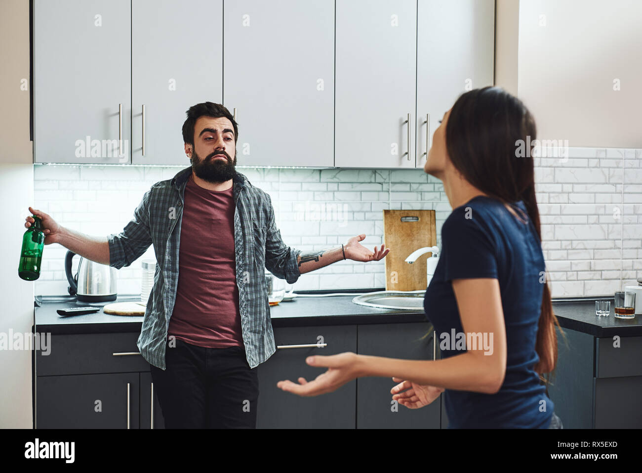 Drunk husband. Dark-haired addicted woman feeling despair while arguing with her drunk husband. He is standing in the kitchen, with a bottle of alcoho Stock Photo