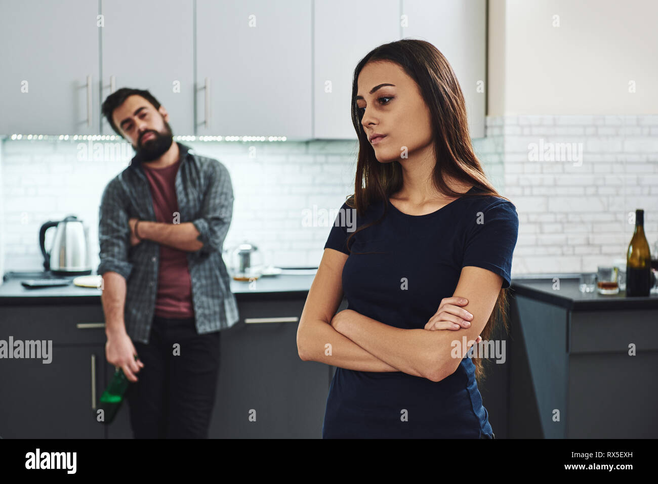 Dark-haired addicted woman feeling despair while standing in the kitchen, thinking about her drunk husband with a bottle of alcohol. He is looking at  Stock Photo
