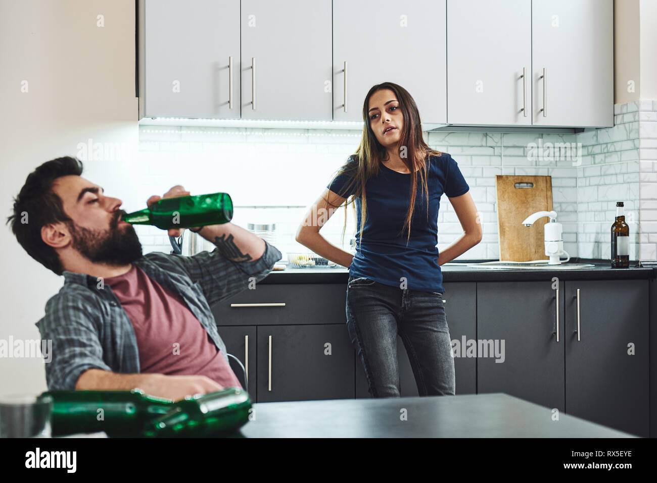 Drunk husband. Dark-haired addicted woman feeling despair while looking at her drunk husband. He is drinking in the kitchen, wasted. Empty bottles are Stock Photo