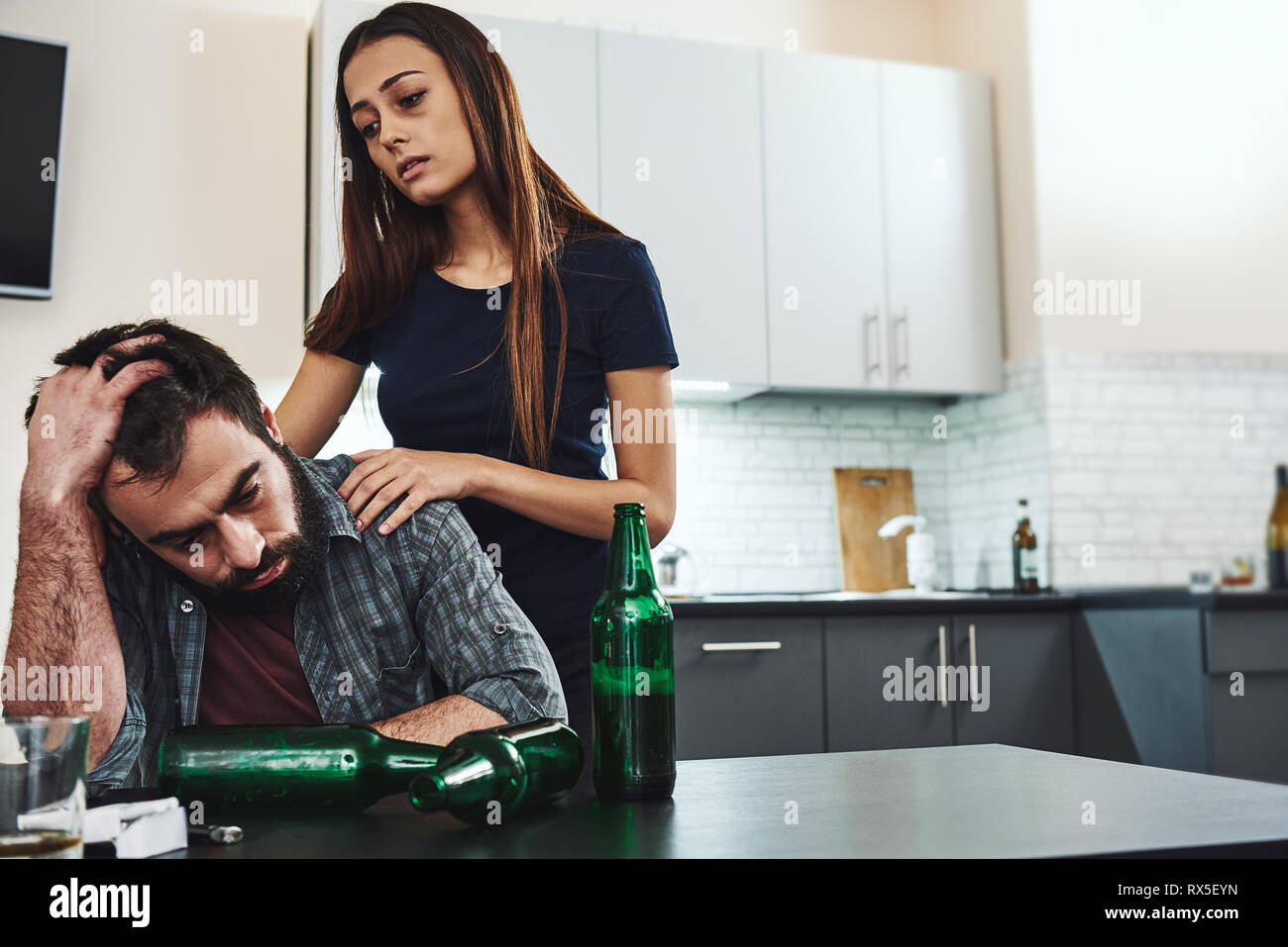 Drunk husband. Dark-haired addicted woman feeling despair while standing near her drunk husband. He is sitting in the kitchen, wasted. Empty bottles a Stock Photo