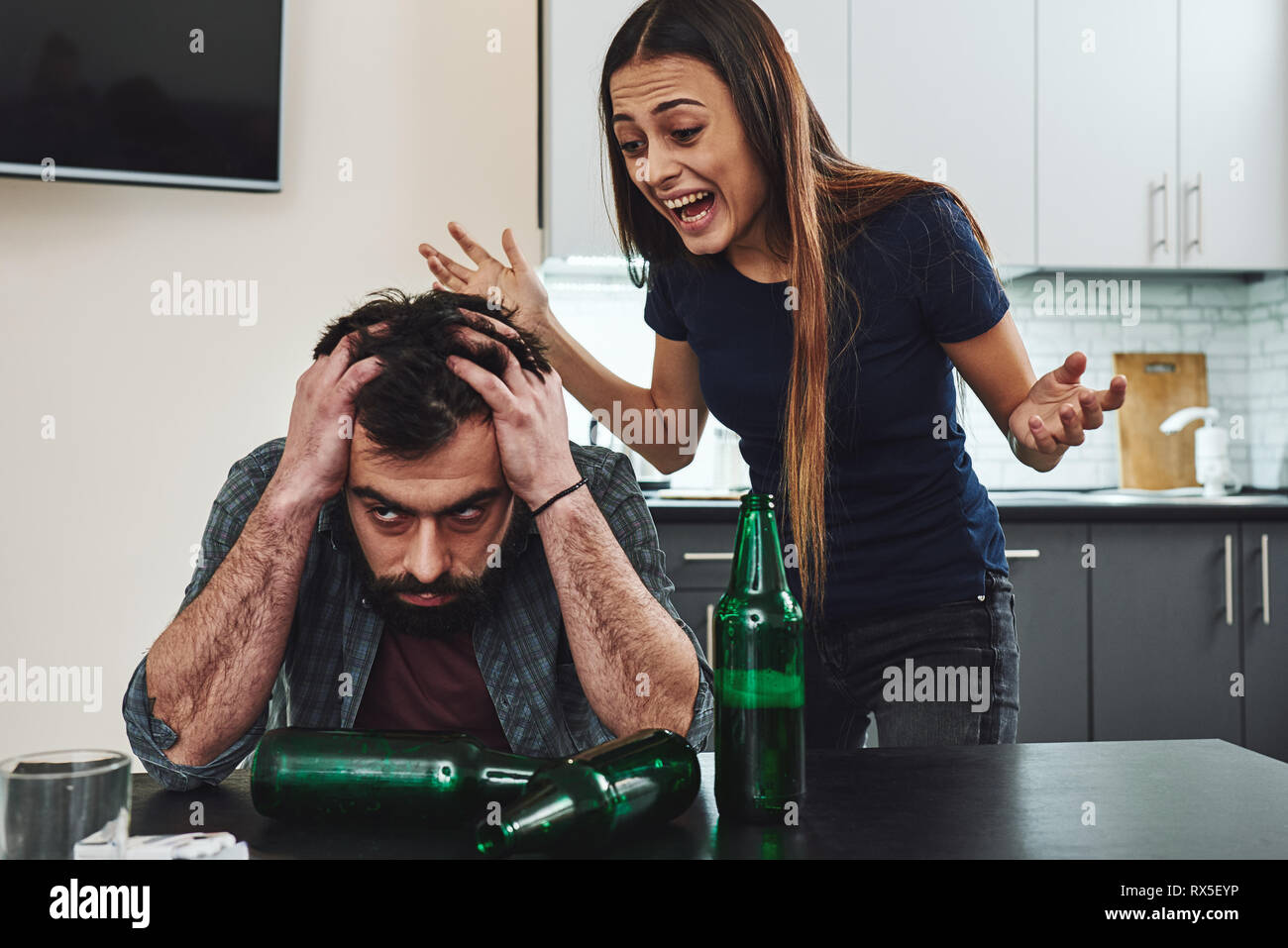 Drunk husband. Dark-haired addicted woman feeling despair while shouting at her drunk husband sitting in the kitchen. Empty bottles are on the table Stock Photo