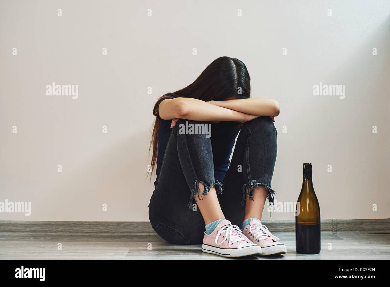 Sad dark-haired woman sitting on the floor hugging her knees with a bottle of red wine crying, divorce concept, parting, alcoholism. Horizontal shot Stock Photo