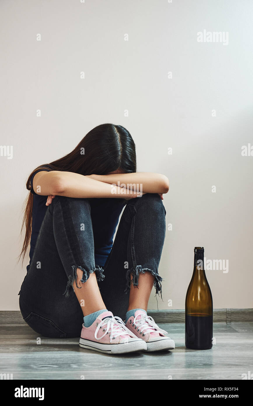 Sad dark-haired woman sitting on the floor hugging her knees with a bottle of red wine crying, divorce concept, parting, alcoholism. Vertical shot Stock Photo