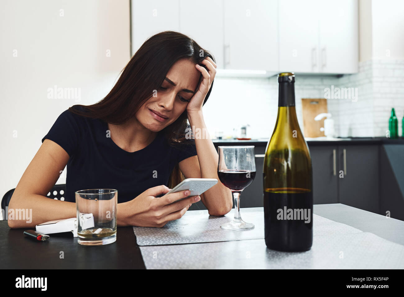 Portrait of young woman in despair, holding her phone and crying while sitting at table, drinking alcohol at home. She is waiting for phone call with  Stock Photo