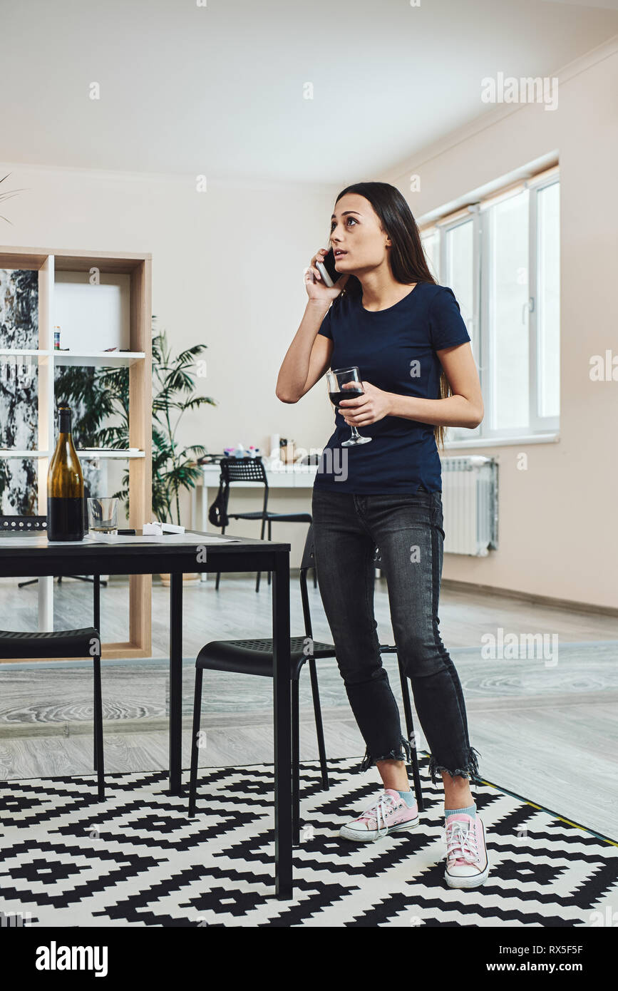 Dark-haired, sad and wasted alcoholic woman standing in the kitchen, holding a glass of wine, while talking on the phone at home. She is completely dr Stock Photo