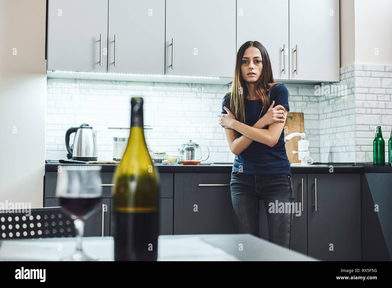 Dark-haired, sad and wasted alcoholic woman standing in the kitchen, looking at red wine on the table, completely drunk, looking depressed, lonely and Stock Photo