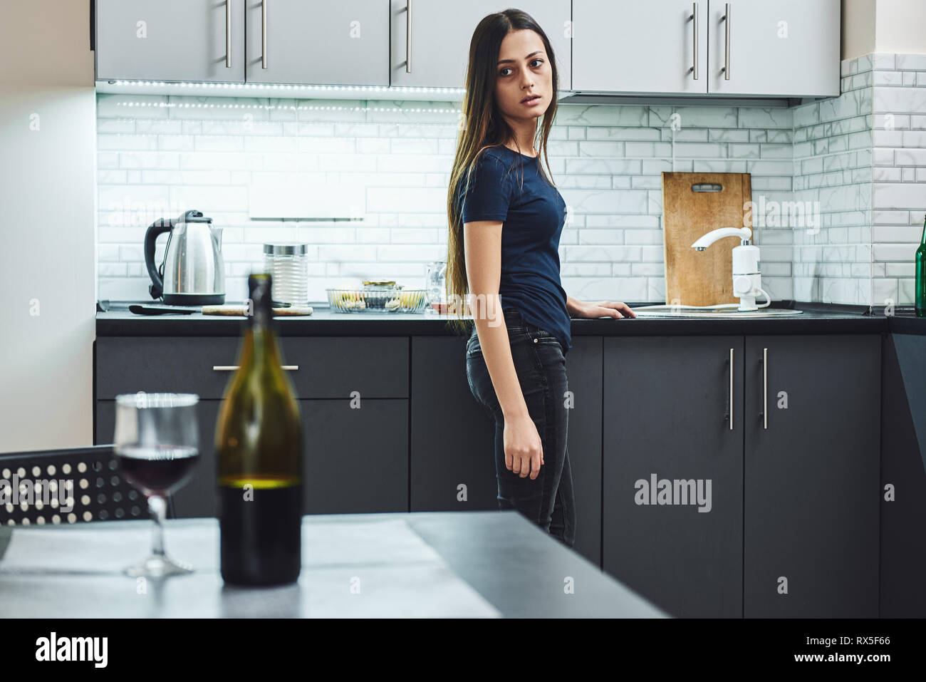 Dark-haired, sad and wasted alcoholic woman standing in the kitchen, looking at red wine on the table, completely drunk, looking depressed, lonely and Stock Photo