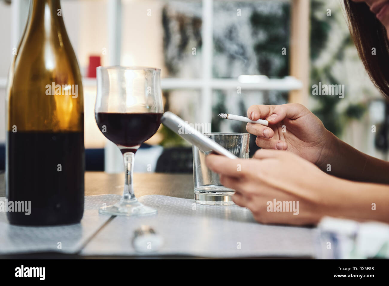 Woman holds a cigarette in one hand, a phone in another. Using of empty glass as an ashtray. A bottle and glass of red wine are on the table. Stock Photo