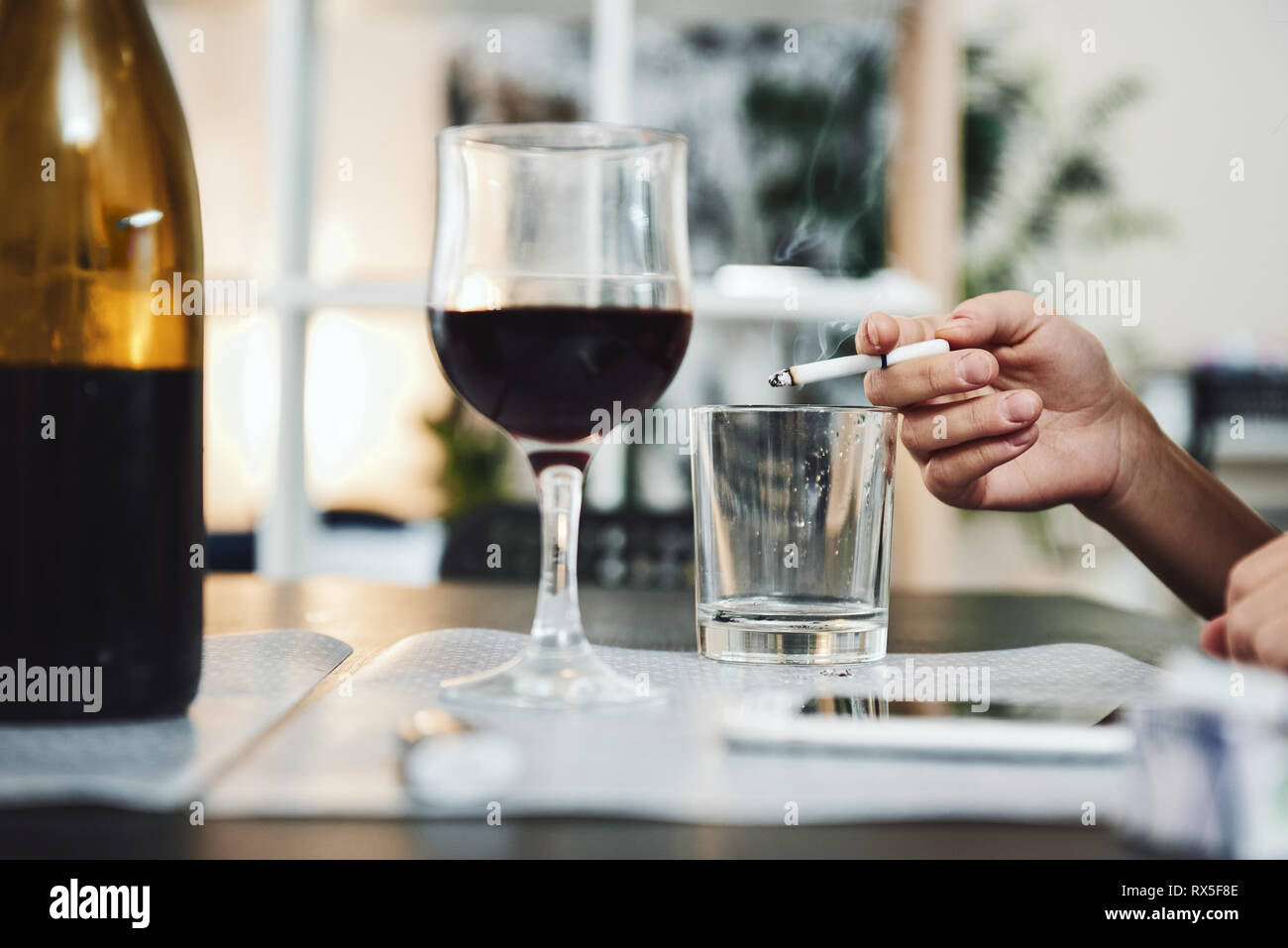 Hand holding a cigarette. Using of empty glass as an ashtray. A bottle and glass of red wine are on the table. Stock Photo