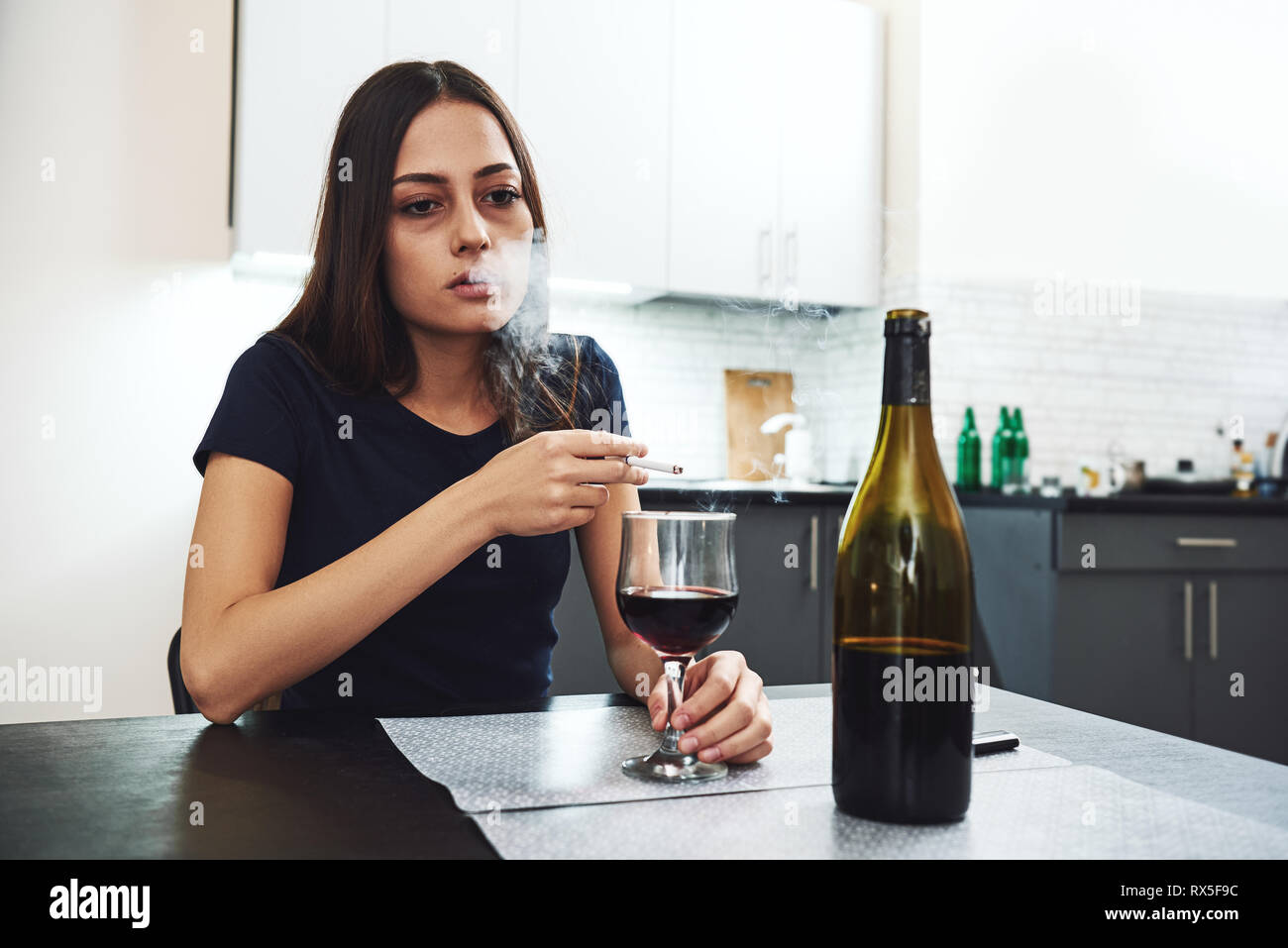 Portrait of young woman in despair, smoking cigarette while sitting at table with glass of alcohol at home. Female alcoholism concept. Protest in the  Stock Photo