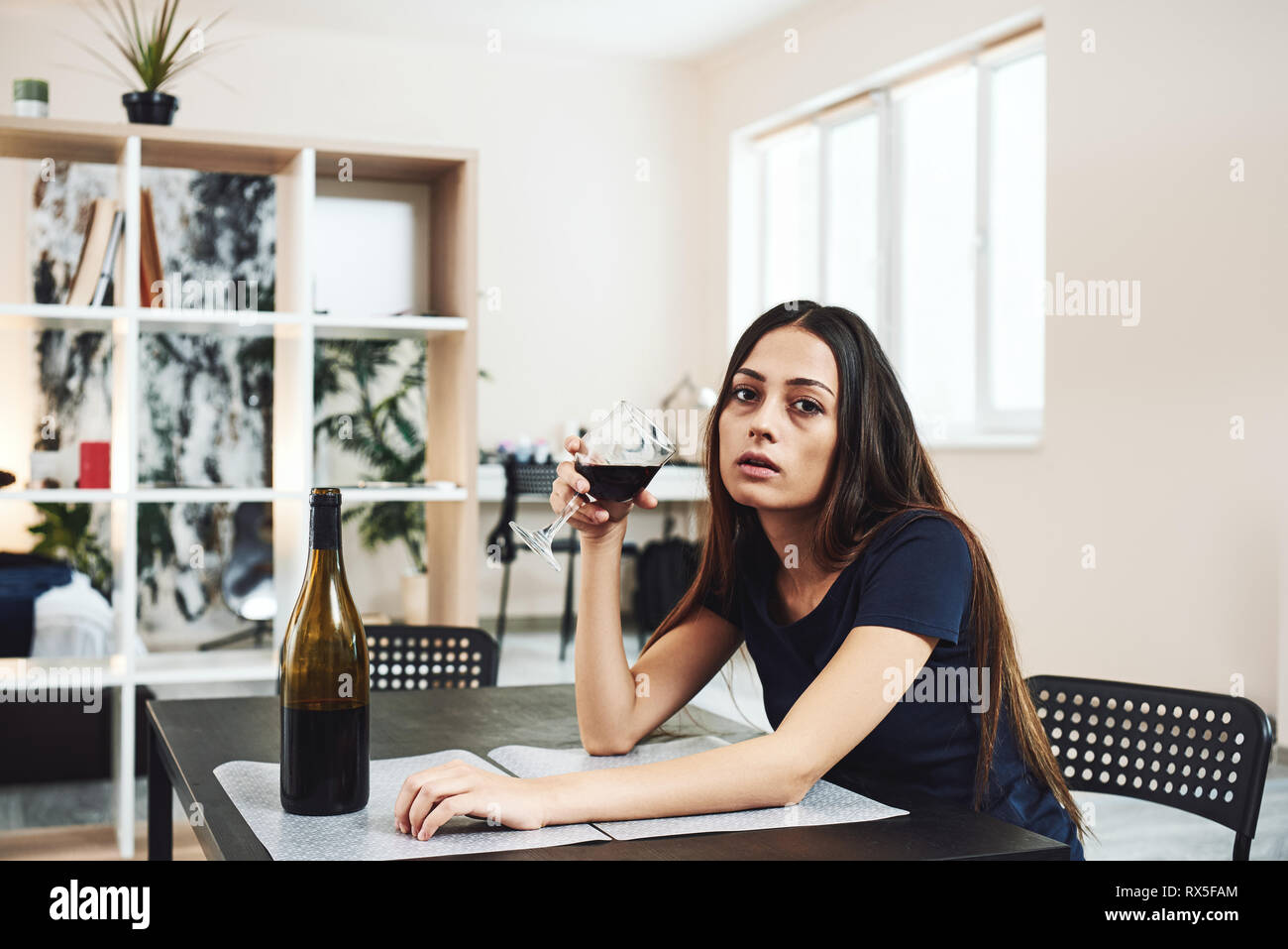 Depressed, divorced woman sitting alone in kitchen at home and holding a glass of red wine because of problems at work and troubles in relationships.  Stock Photo