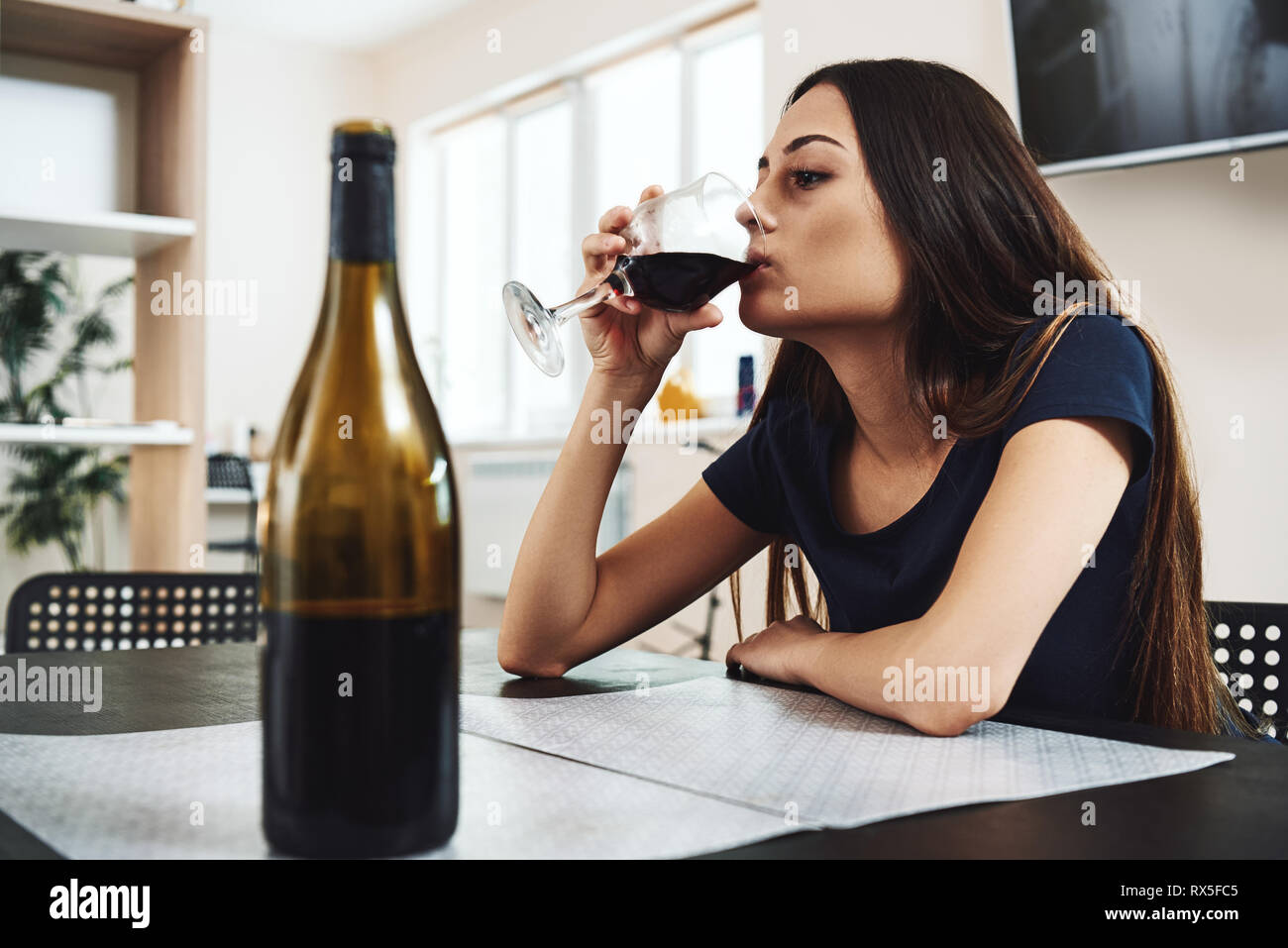 Depressed, divorced woman sitting alone in kitchen at home and drinking a glass of red wine because of problems at work and troubles in relationships. Stock Photo