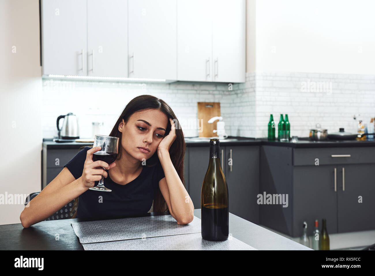 Dark-haired, sad and wasted alcoholic woman sitting at home, in the kitchen, leaning over the table, holding a glass and looking at bottle of red wine Stock Photo