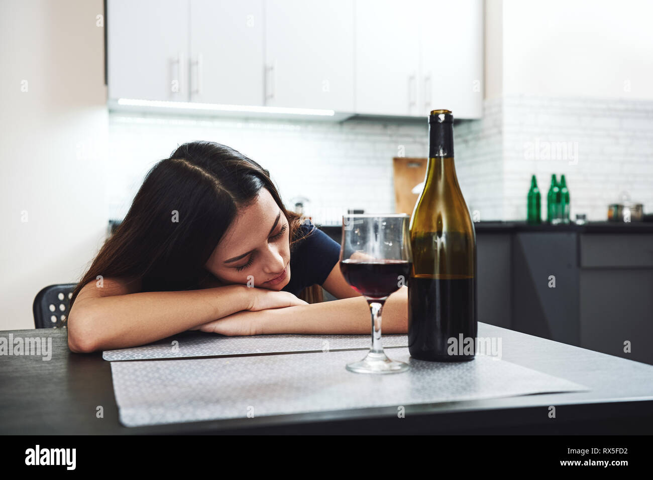 After drinking. Sleepy drunk young woman lying on the table in the kitchen and sleeping with bottle and glass of red wine standing on the table in fro Stock Photo