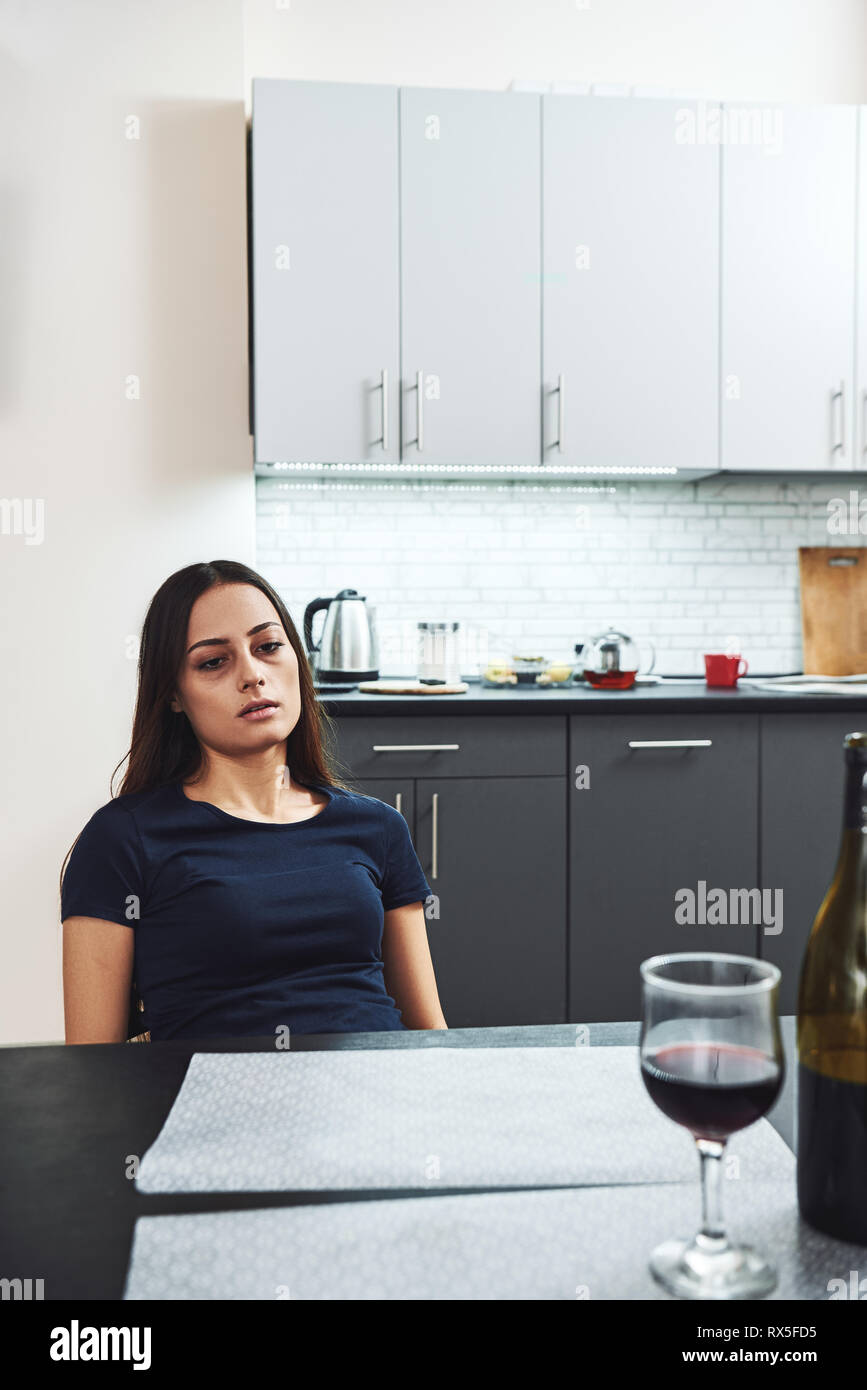 Dark-haired, sad and wasted alcoholic woman sitting at home, in the kitchen, looking at bottle and glass of red wine on the table, completely drunk, l Stock Photo