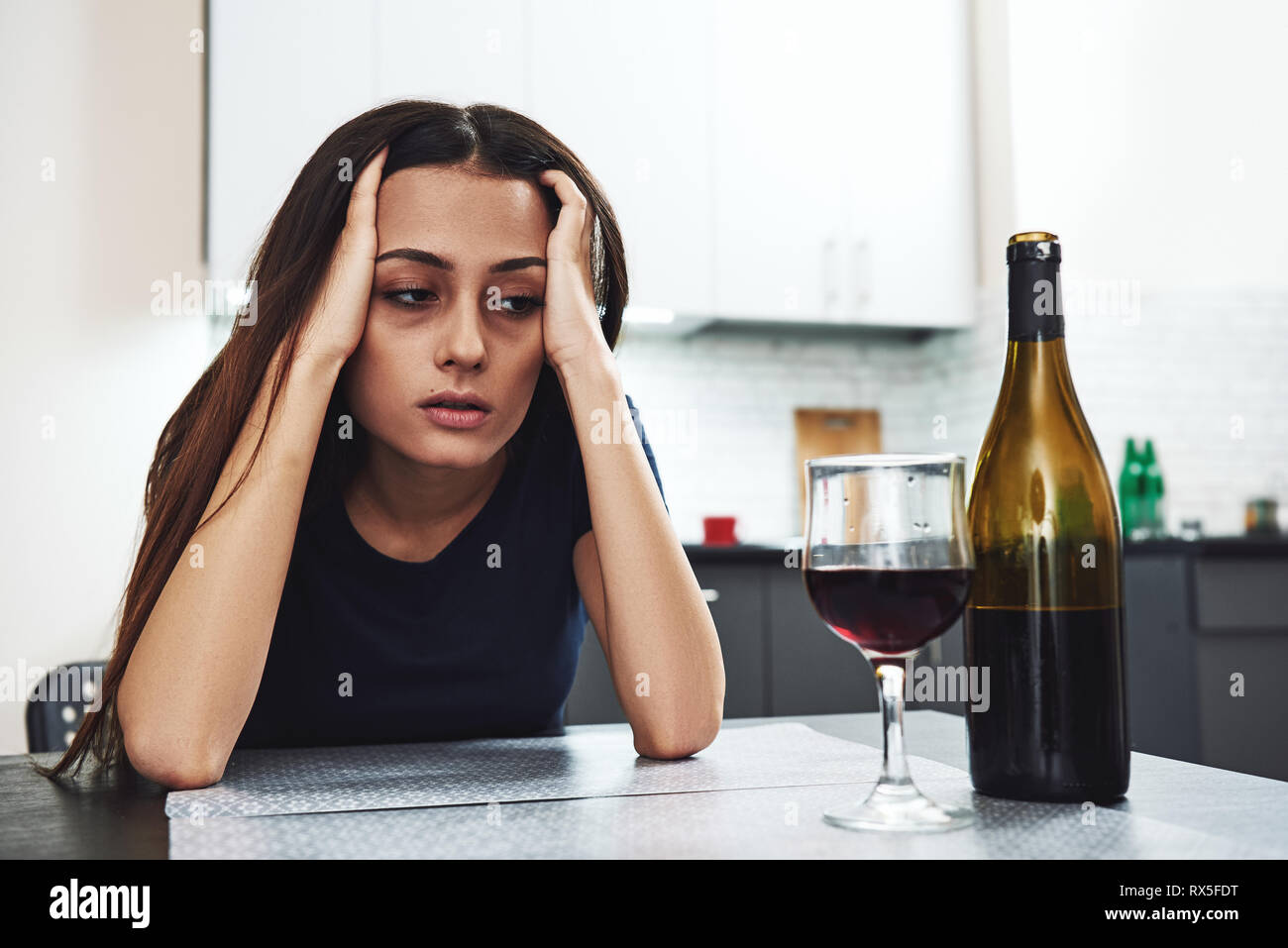 Dark-haired, sad and wasted alcoholic woman sitting at home, in the kitchen, leaning over the table, looking at bottle and glass of red wine on the ta Stock Photo