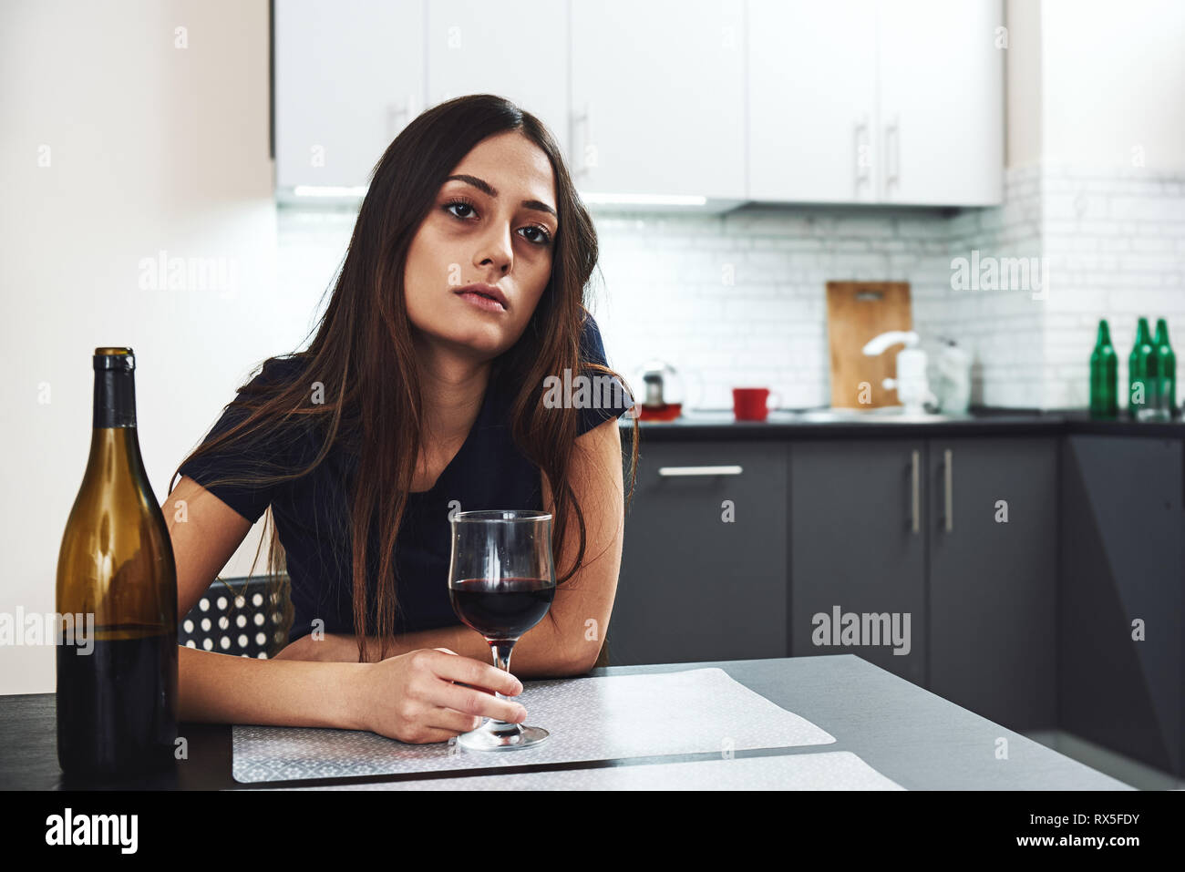 Dark-haired, sad and wasted alcoholic woman sitting at home, in the kitchen, drinking red wine, holding glass, completely drunk, looking depressed, lo Stock Photo