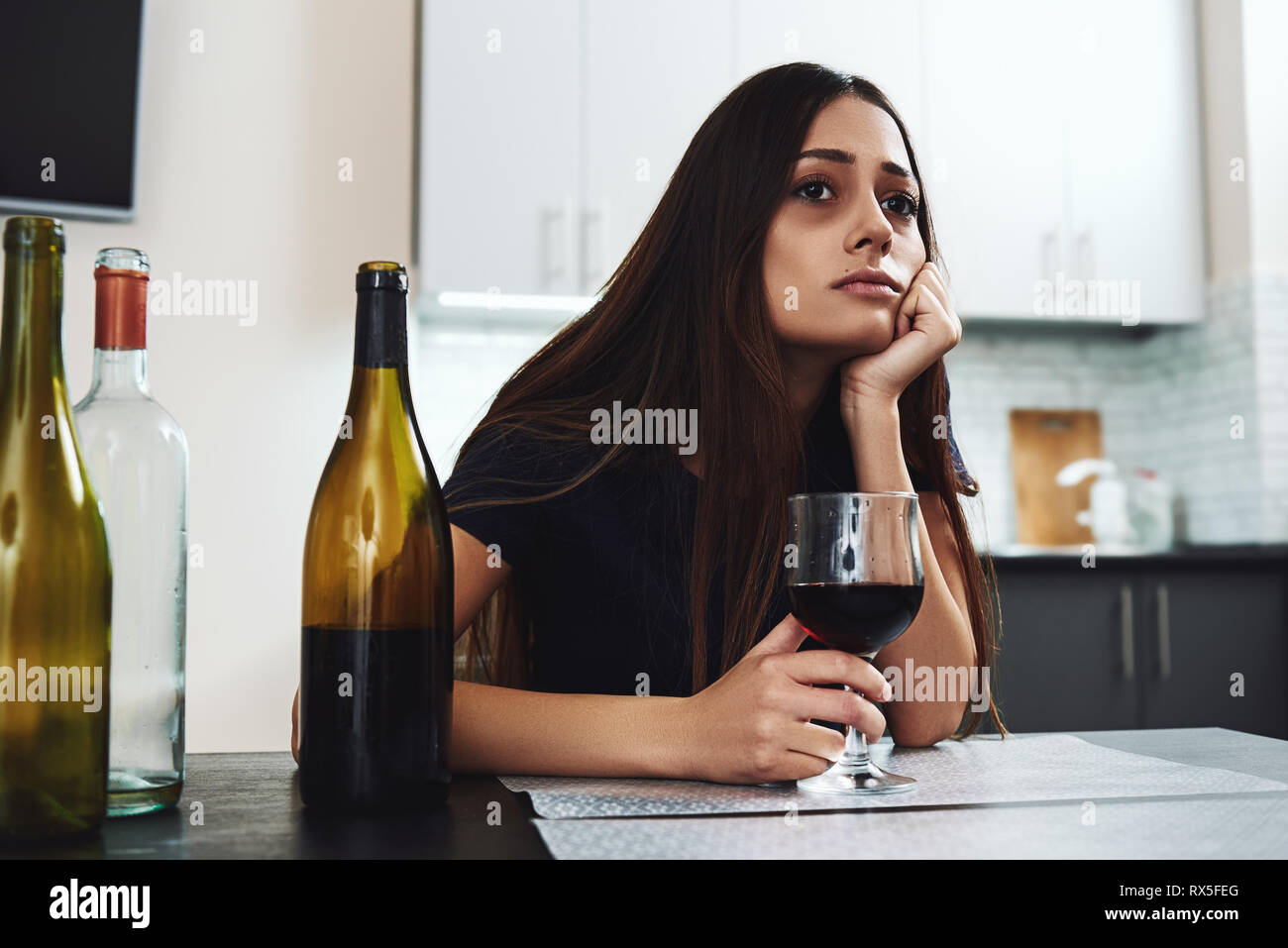 Dark-haired, sad and wasted alcoholic woman sitting at home, in the kitchen, drinking red wine, holding glass, completely drunk, looking depressed, lo Stock Photo