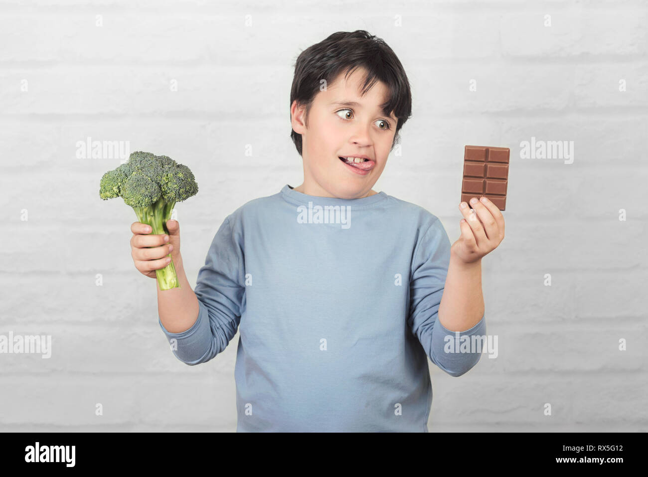 Hungry child with broccoli and an chocolate bar in his hands against brick background Stock Photo