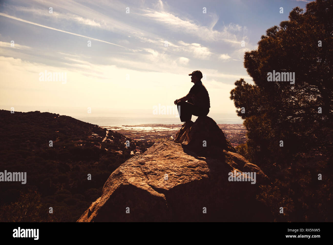 Man with peaked cap sitting on a rock on the top of a mountain, resting and enjoying the view Stock Photo