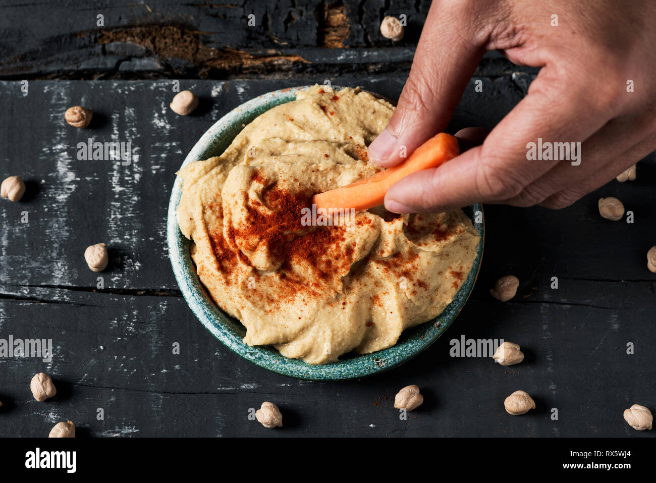 high angle view of a man dipping a strip of carrot in a homemade hummus seasoned with paprika served in a green ceramic plate, on a dark gray rustic w Stock Photo