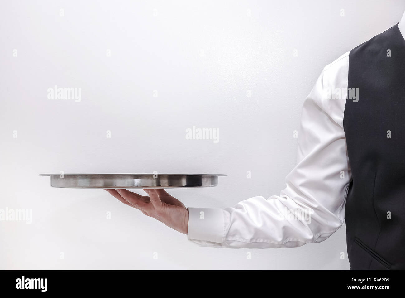 A waiter / butler in white shirt and black suit vest carrying an empty silver tray. Copy space on white background. Stock Photo