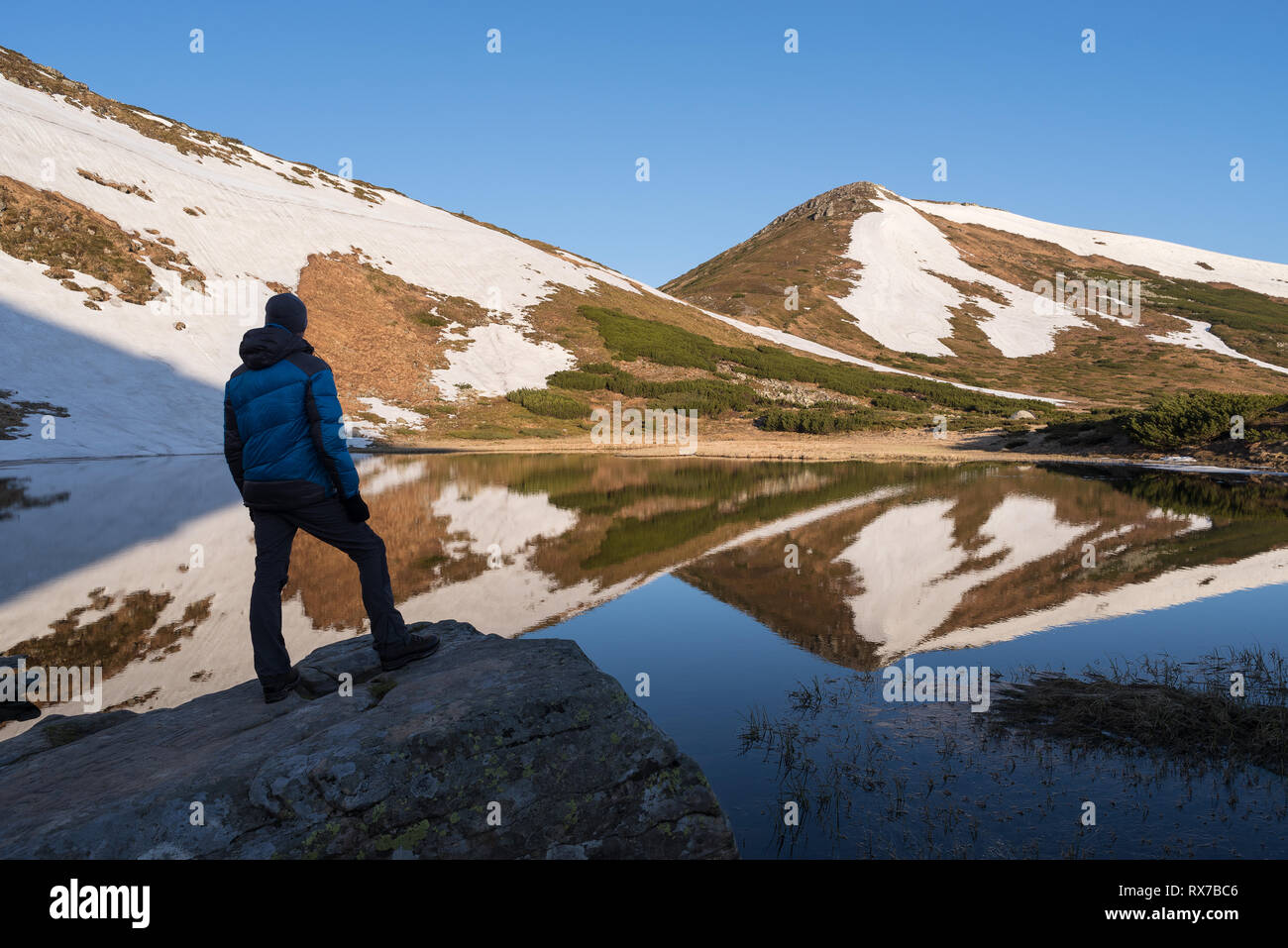 Spring landscape with a mountain lake. Man tourist is standing on a stone and looking at the top of the mountain. Sunny morning with blue sky Stock Photo