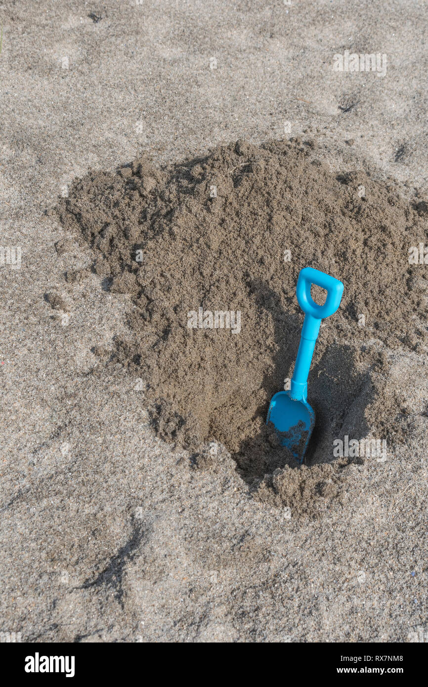 Seaside hole dug into sand, with kids blue spade. Metaphor 'dig a hole