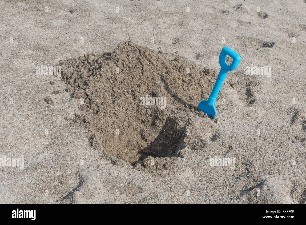 Seaside hole dug into sand, with kids blue spade. Metaphor 'dig a hole