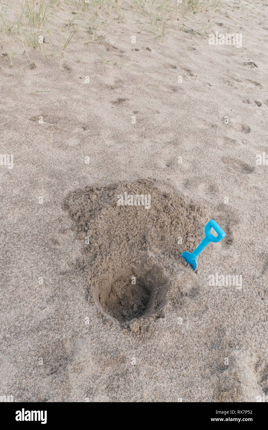 Seaside hole dug into sand, with kids blue spade. Metaphor 'dig a hole