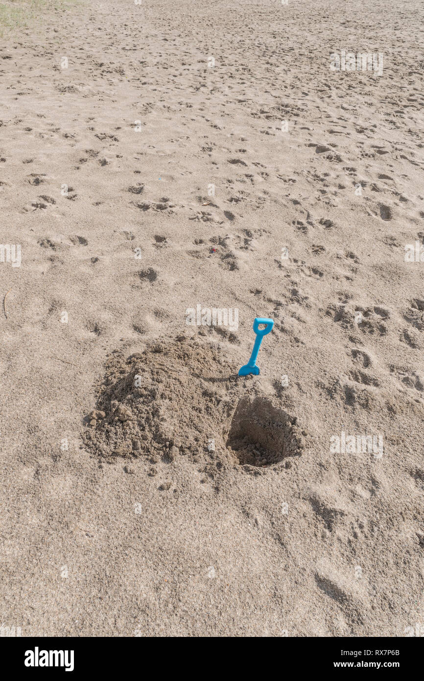 Seaside hole dug into sand, with kids blue spade. Metaphor 'dig a hole