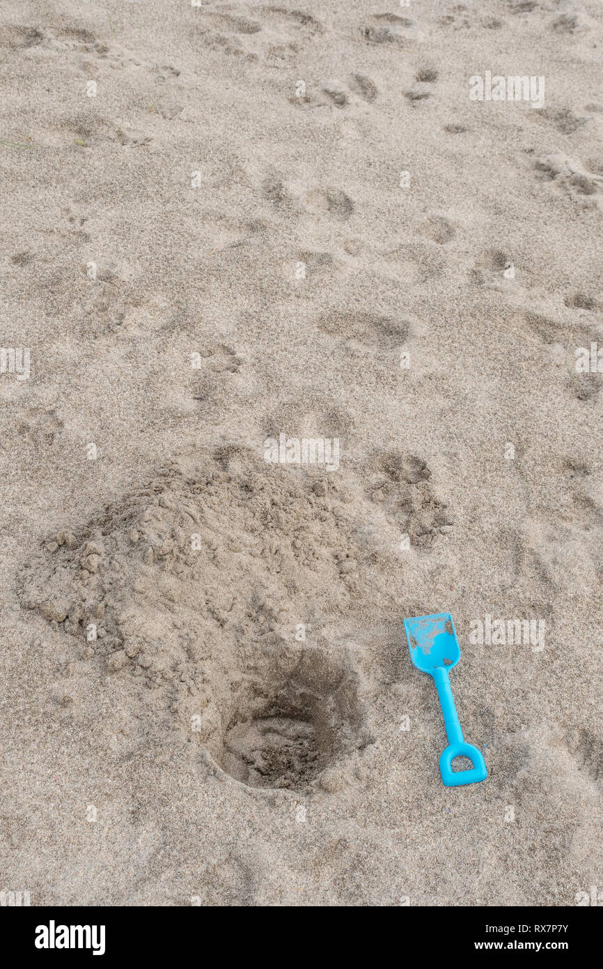 Seaside hole dug into sand, with kids blue spade. Metaphor 'dig a hole