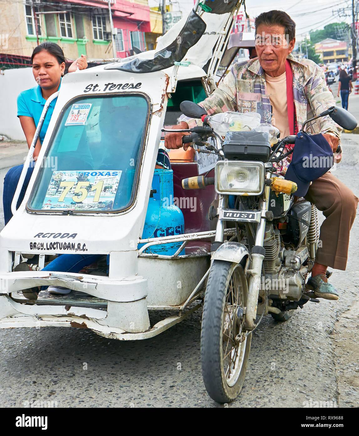 Roxas City, Capiz Province, Philippines: Low-angle view of a passing old tricycle with a male driver and female passenger and cargo Stock Photo