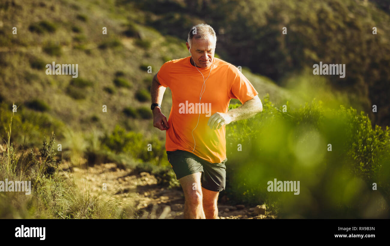Senior man running on a rough hilly terrain on a sunny day. Man trail running on a hill listening to music. Stock Photo