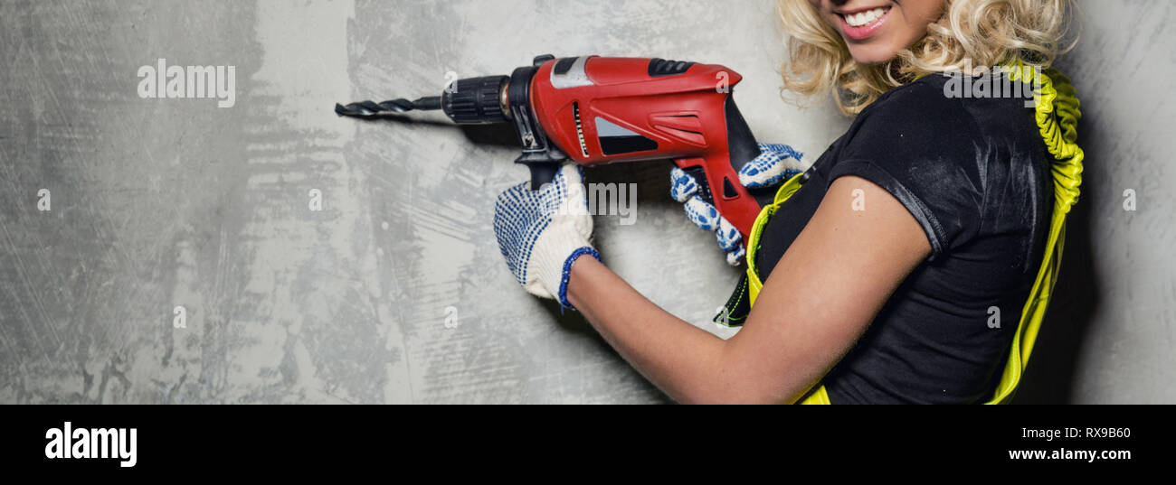 Attractive builder woman wearing protective coverall workwear smile look at camera holding a drill in her hands pose over obsolete horizontal wall Stock Photo