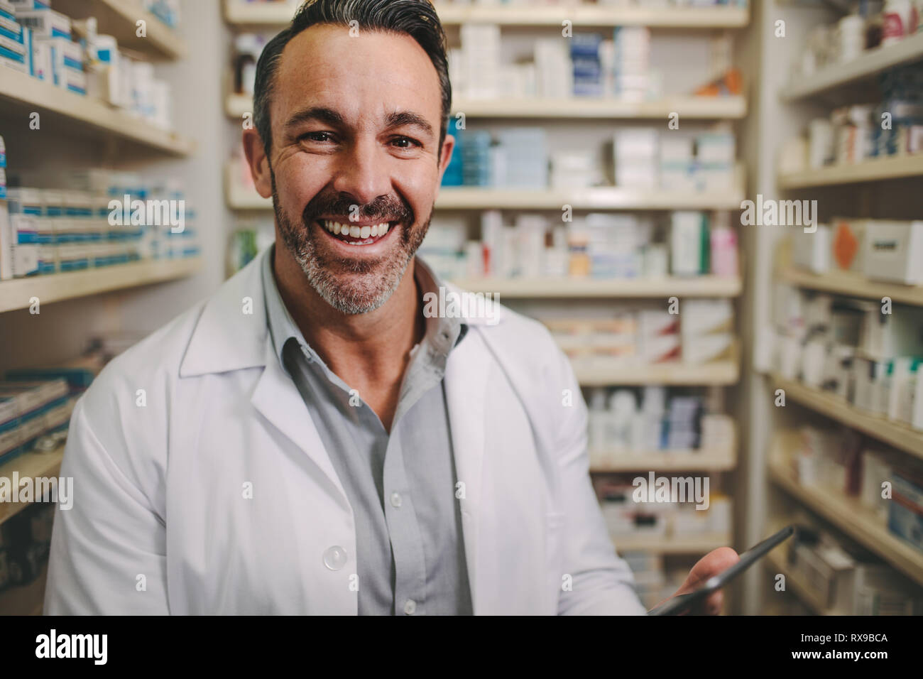 Smiling mature pharmacist with digital tablet standing by aisle in drug store. Male pharmacist