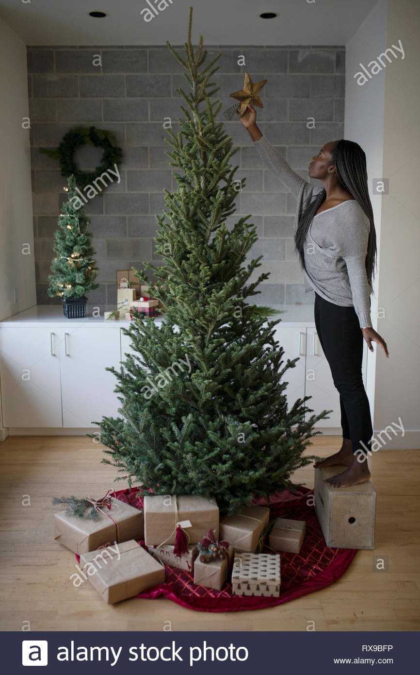 Young woman on box reaching to place star on christmas tree Stock Photo