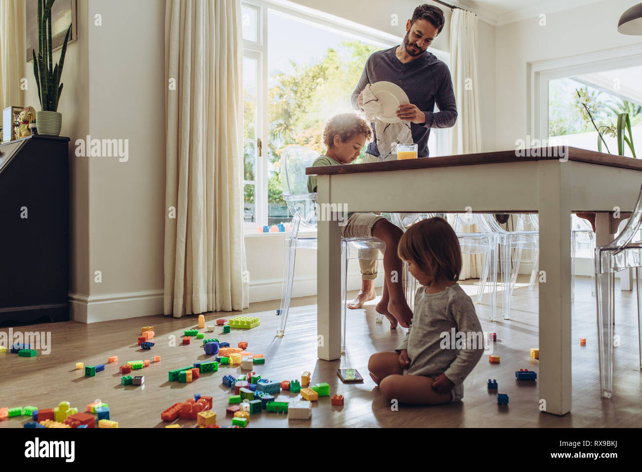 Man looking after his kids while doing the household works. Kid playing with toys sitting on floor with his father doing daily household chores. Stock Photo