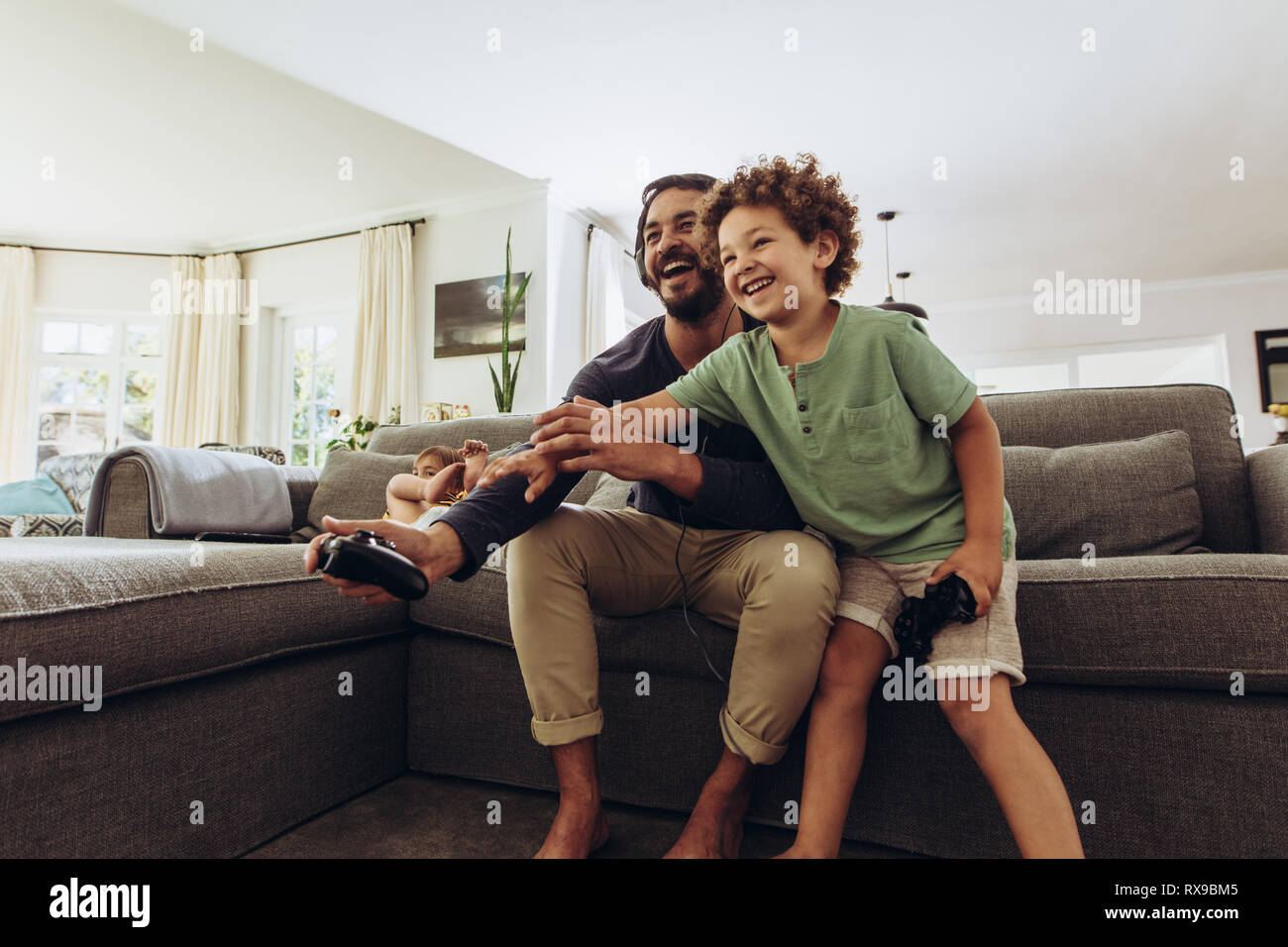 Father and son having fun playing video game at home. Man playing video game with son sitting on couch at home holding joysticks. Stock Photo
