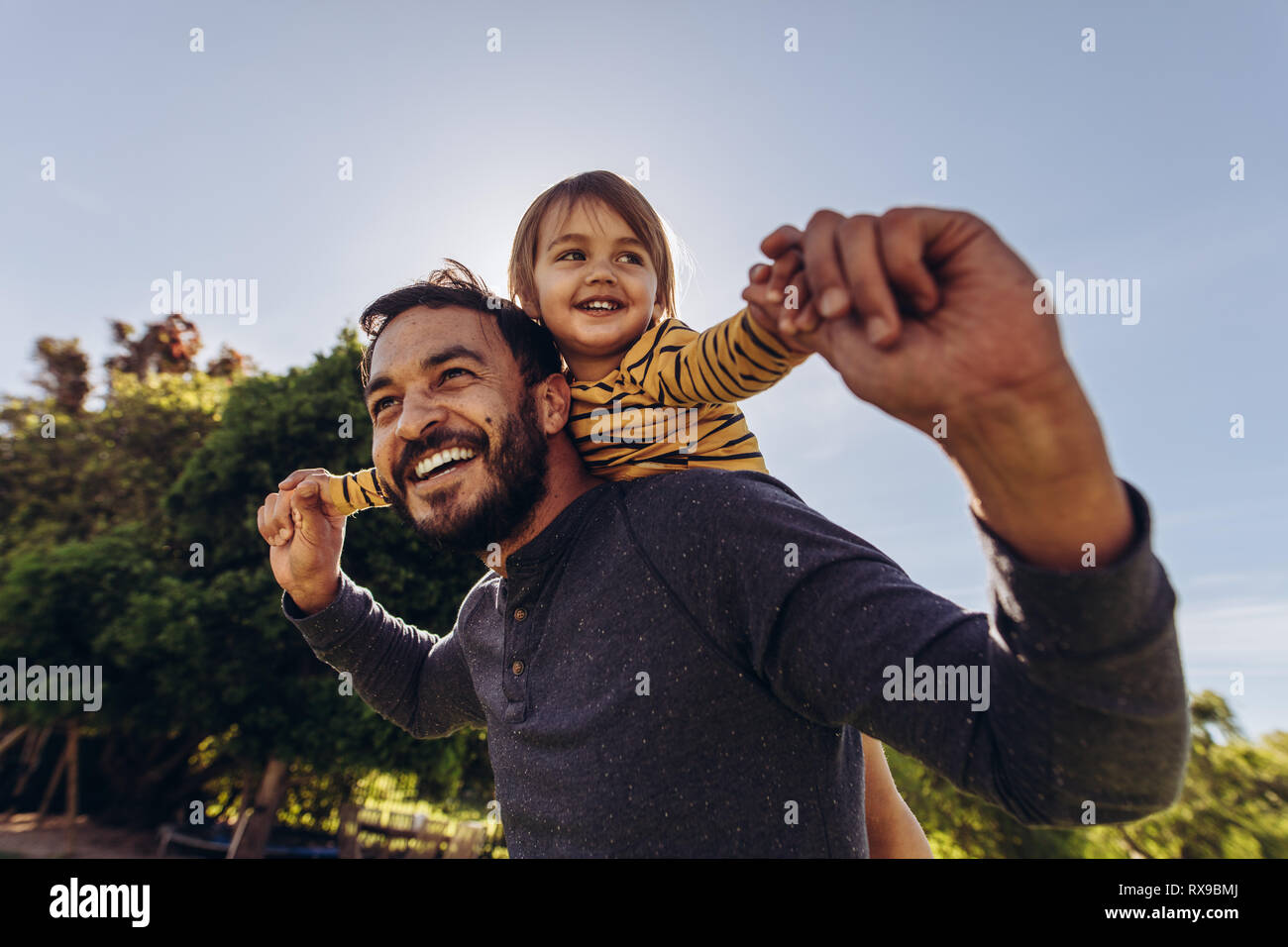 Close up of a man carrying his child on his back outdoors. Happy father walking outdoors with his kid on his back. Stock Photo