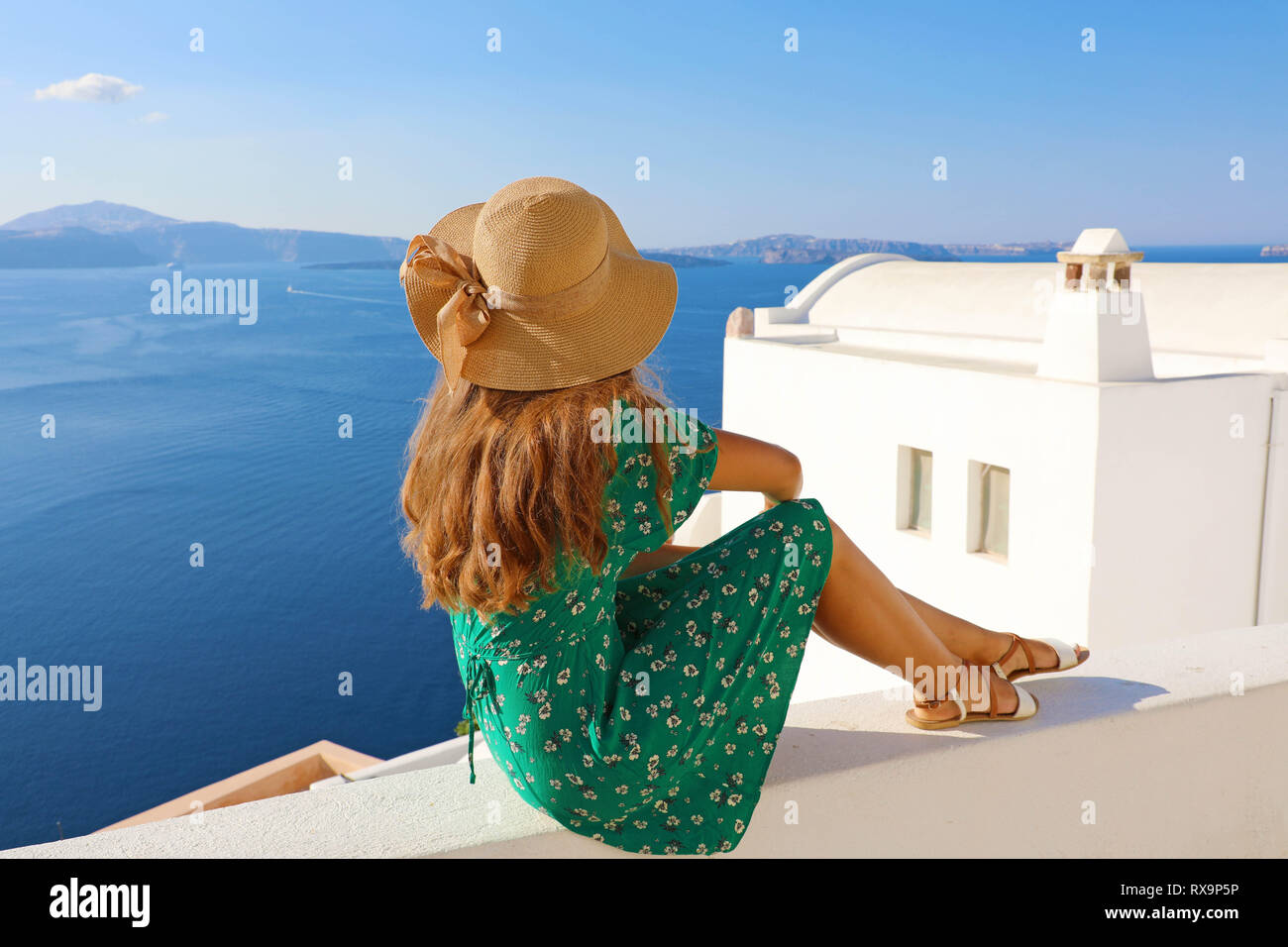 Young woman sitting on wall and looking to beautiful landscape in Santorini, Greece. Rear view. Stock Photo