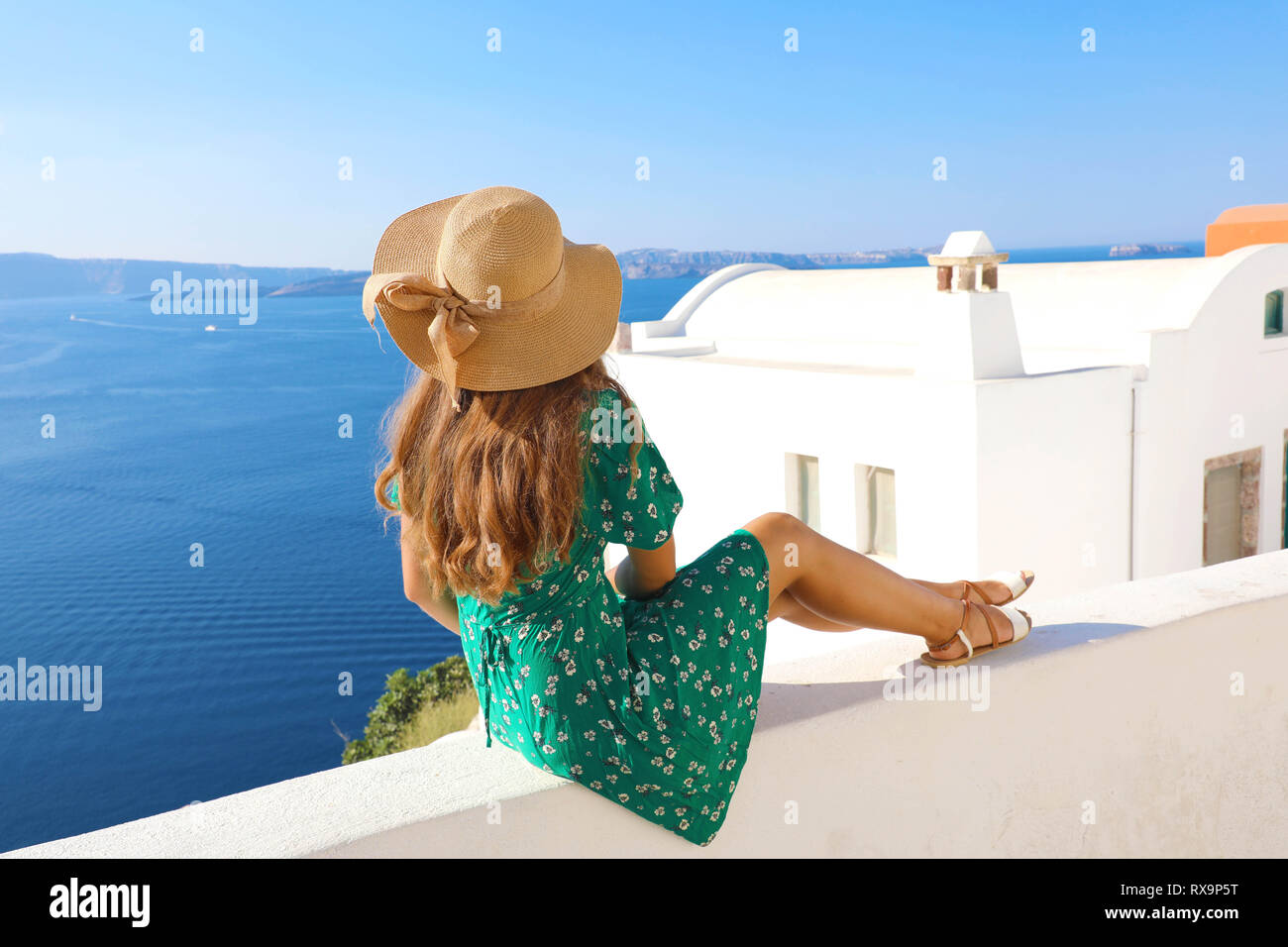 Beautiful young woman sitting on wall looking at stunning view of Mediterranean sea and Santorini village, Greece, Europe. Lifestyle woman with straw  Stock Photo