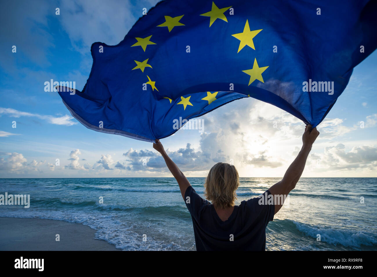 Fluttering European Union flag flying in bright morning sunlight held up by man with blond hair standing on empty Mediterranean beach Stock Photo