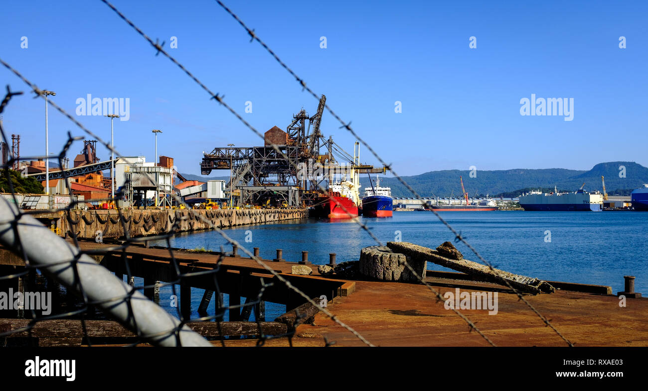 Ships loading coal and iron ore at industrial port taken through razor wire fence in foreground at an Australian Port, NSW. Trade war tariff concept Stock Photo