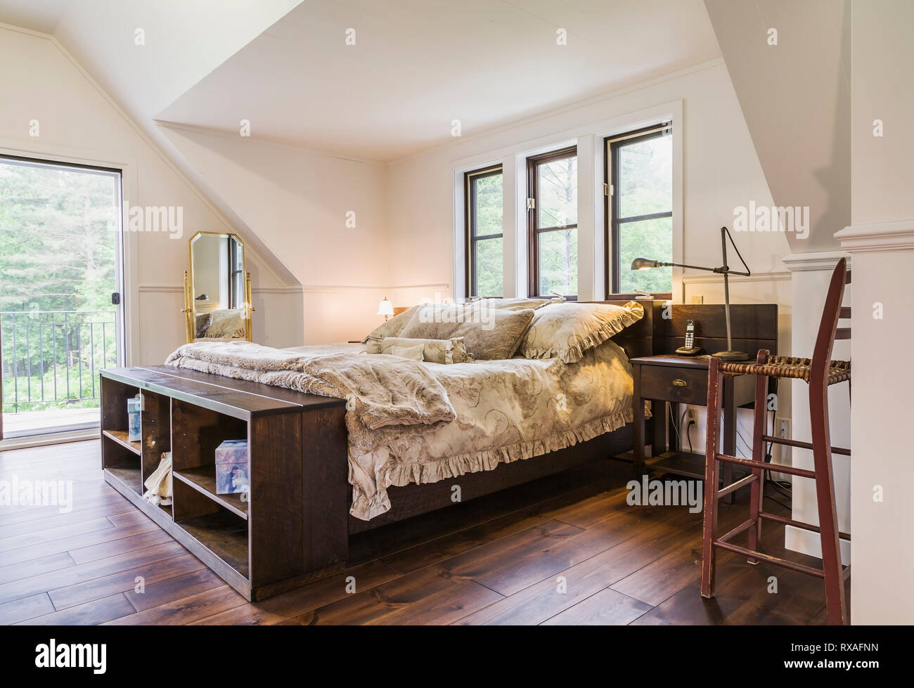 Wood-frame king size bed with flowery bedspread, brass mirror and antique wooden high chair in master bedroom on upper floor inside a New Hampton style home, Quebec, Canada. This image is property released. CUPR0208 Stock Photo