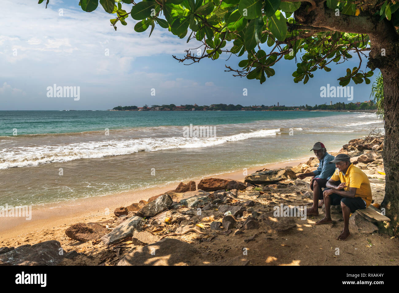 Two men take a break from the heat of the midday sun and chat together beneath the shade of a tree on the beach at Galle in the Southern Province of S Stock Photo