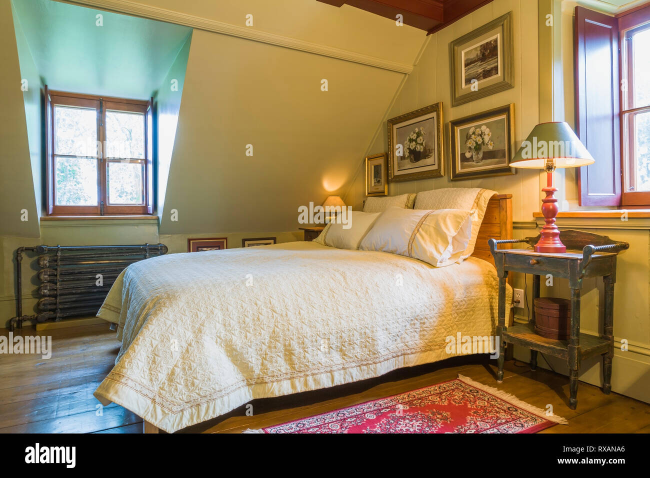 Queen size bed with antique wooden headboard, bedside table and framed paintings in upstairs master bedroom inside an old circa 1805 Canadiana cottage style home, Quebec, Canada. This image is property released. CUPR0323 Stock Photo