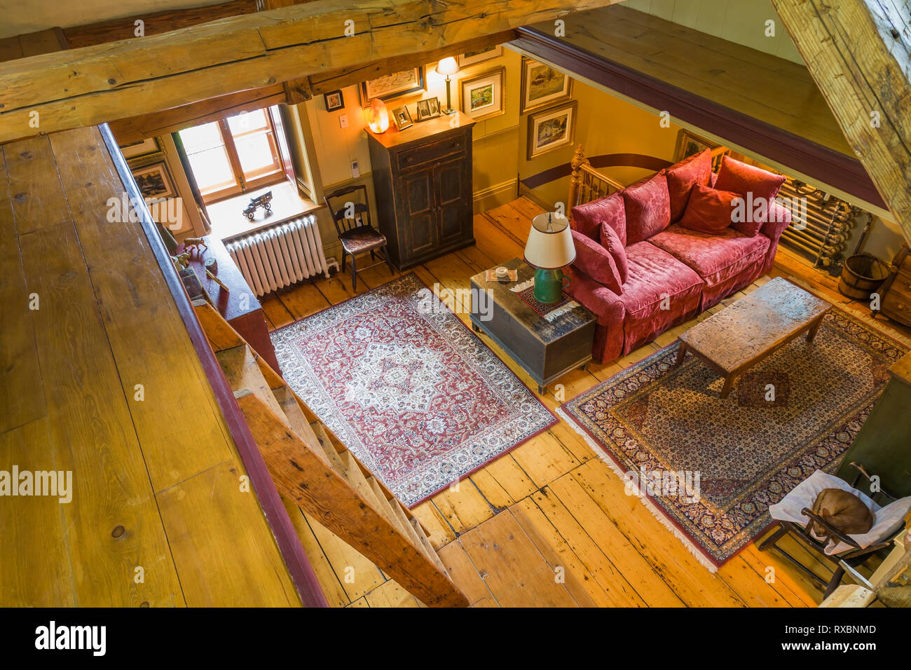 Top view of upstairs family room with wooden Miller's stairs, crimson red upholstered sofa, chest of drawers, armoire and rugs inside an old circa 1805 Canadiana cottage style home, Quebec, Canada. This image is property released. CUPR0323 Stock Photo