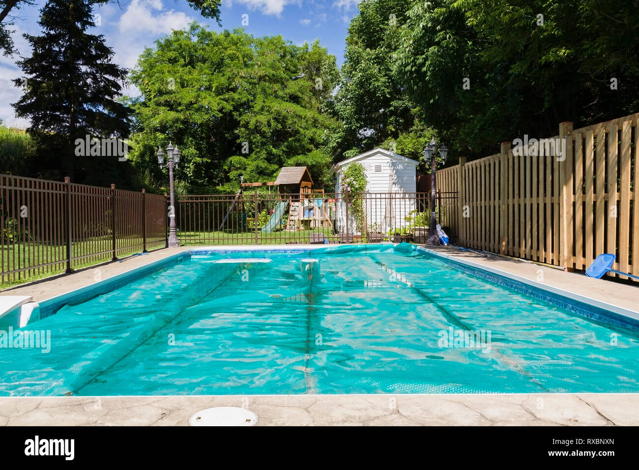 In-ground rectangular swimming pool with protective green vinyl leaf cover enclosed by brown aluminium and wooden security fences in residential backyard in summer, Quebec, Canada. This image is property released. CUPR0329 Stock Photo